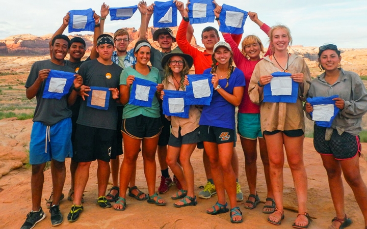 A group of students smile for a group photo. Each person is holding a small blue peter flag.